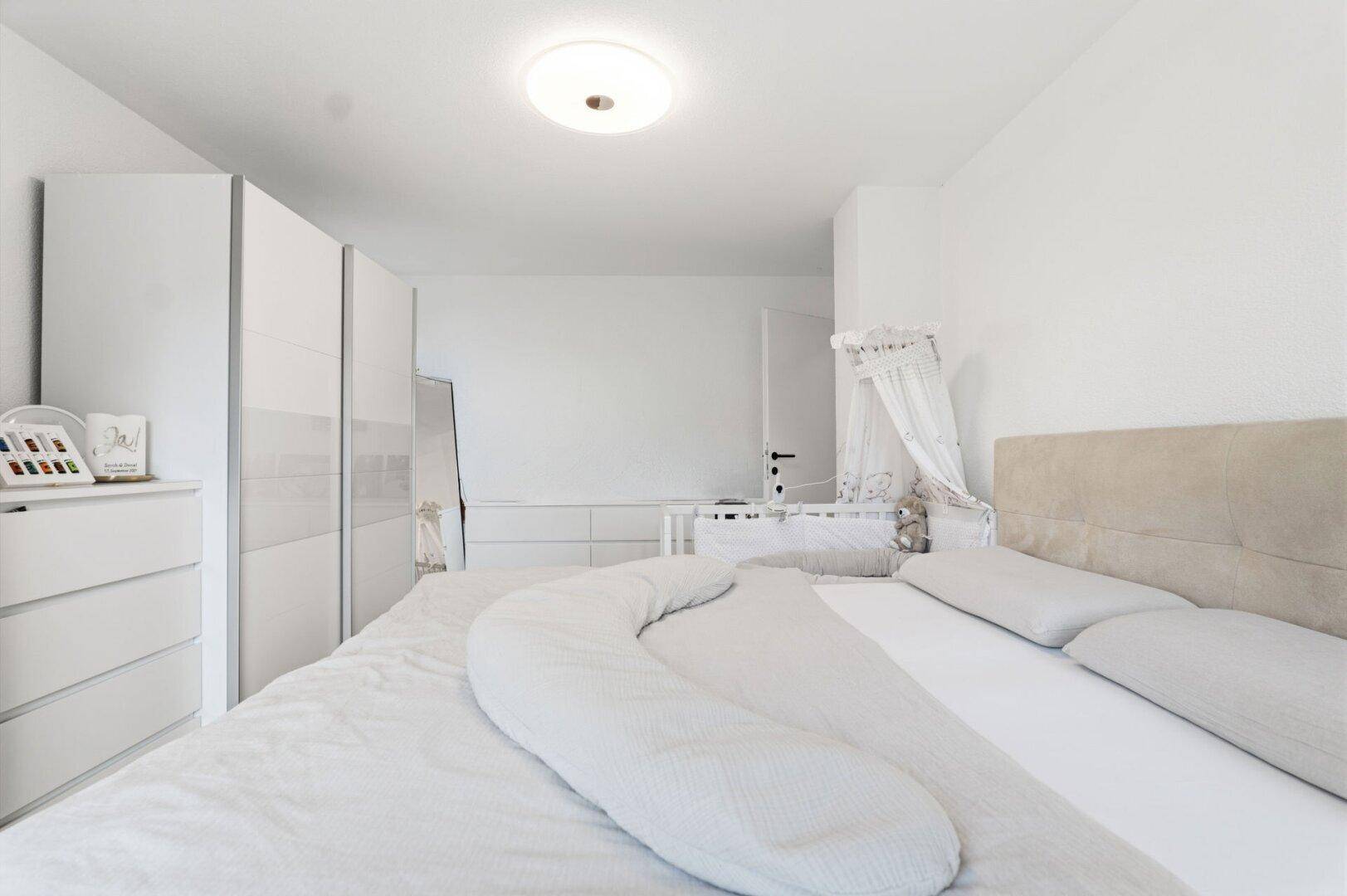 Helles Schlafzimmer mit großem Bett, weißen Schränken und Babybett.