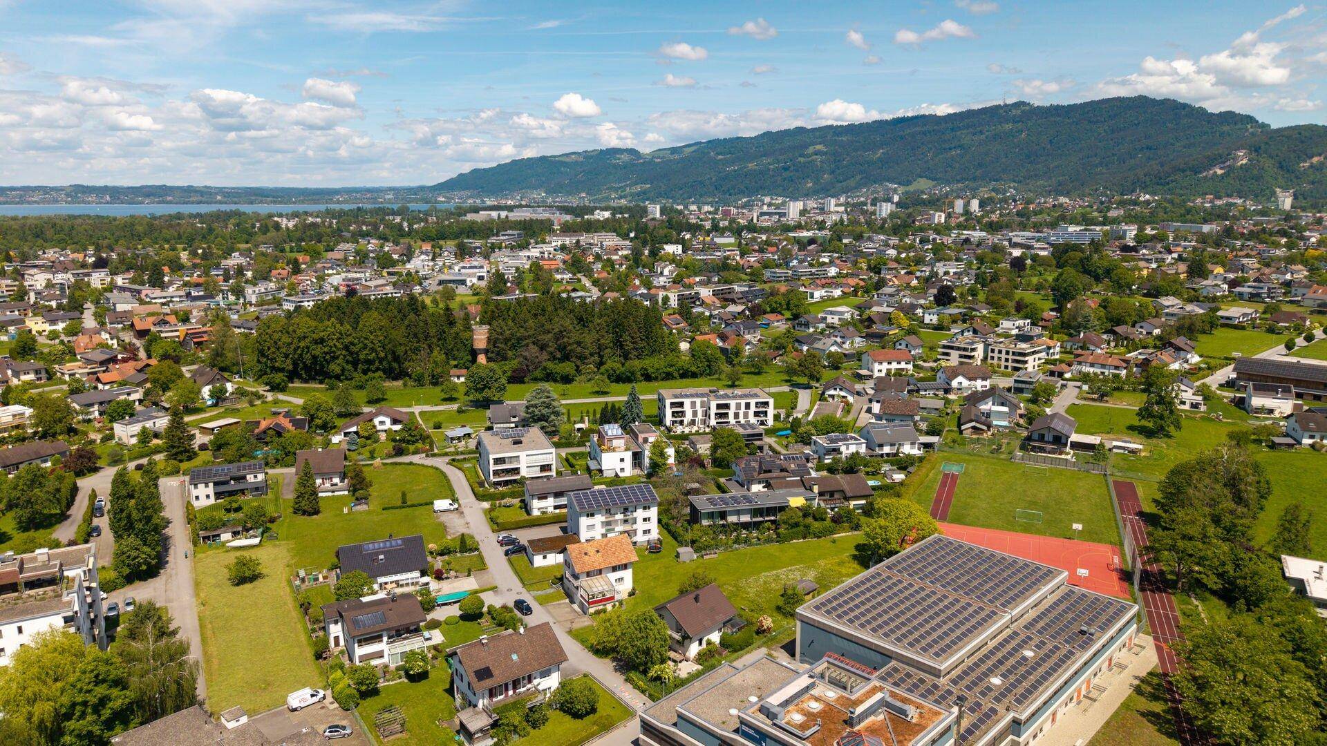 Luftaufnahme einer Wohngegend mit Sportplatz, Bergen im Hintergrund und weitem Blick über die Stadt.