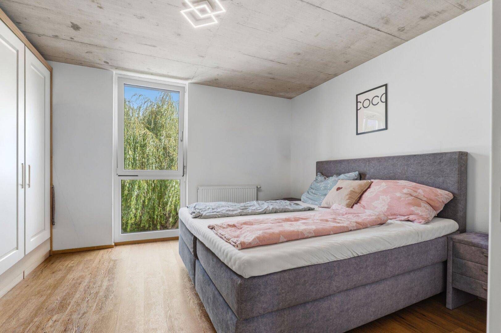 Helles Schlafzimmer mit modernem Bett, weißem Kleiderschrank und Blick ins Grüne durch das Fenster.
