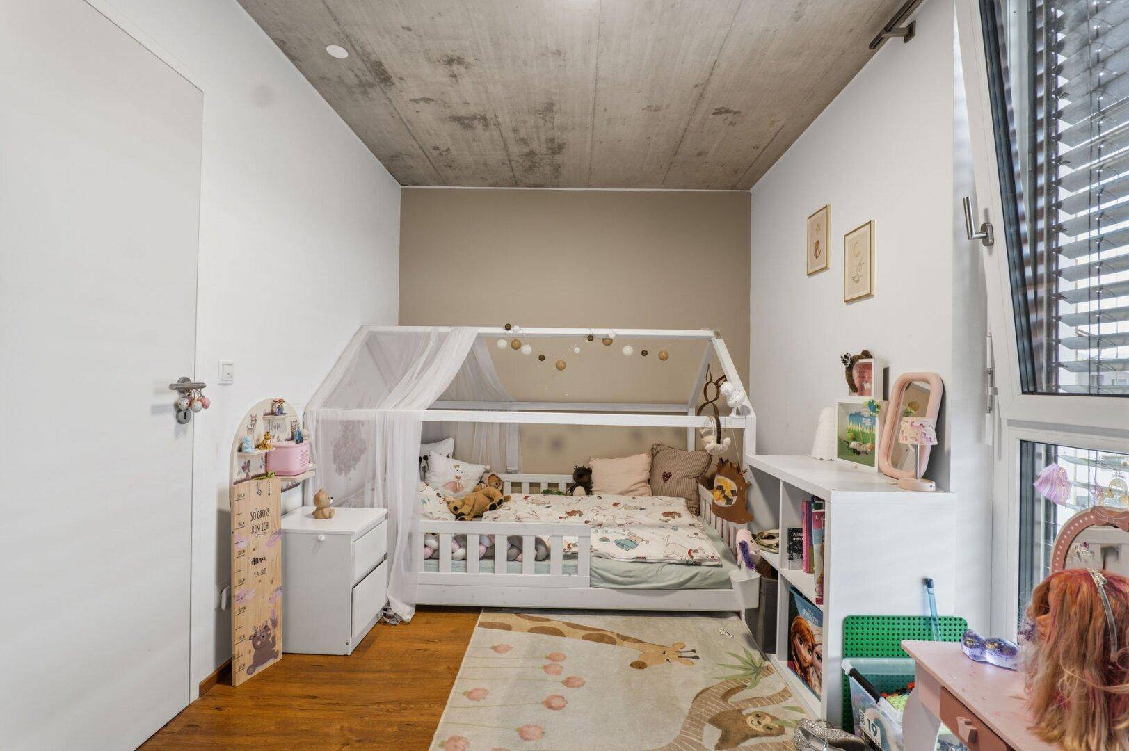 Freundliches Kinderzimmer mit Hausbett, Spielzeug und viel Tageslicht durch das Fenster.