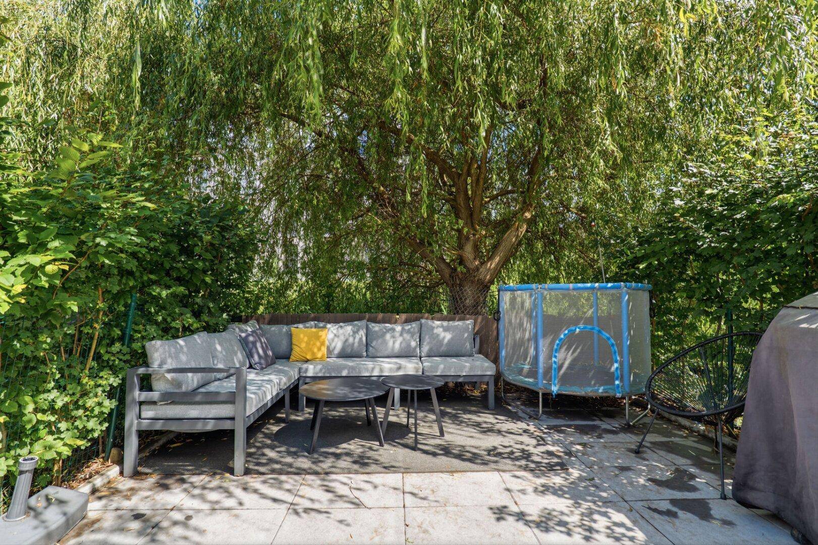 Entspannter Gartenbereich mit Lounge-Sofa, Couchtisch und kleinem Trampolin unter einem Baum.