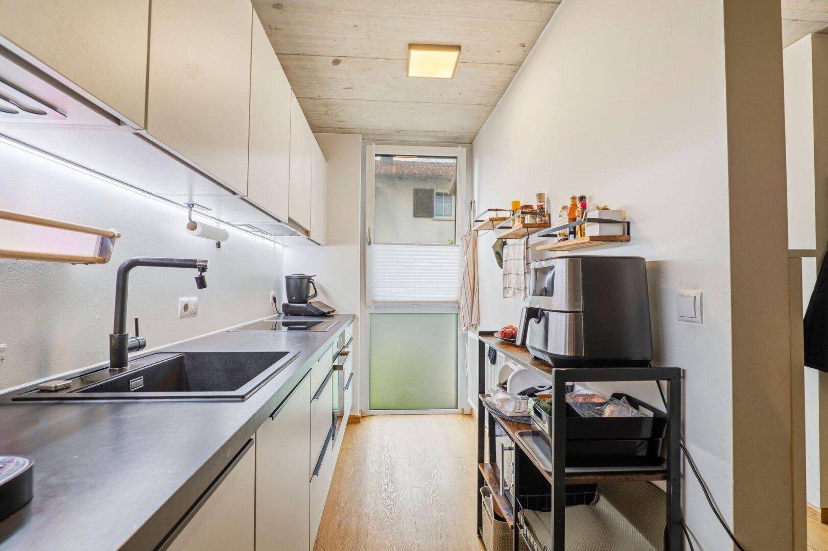 Schmale, moderne Küche mit weißen Schränken, dunkler Arbeitsplatte und Fenster für Tageslicht.