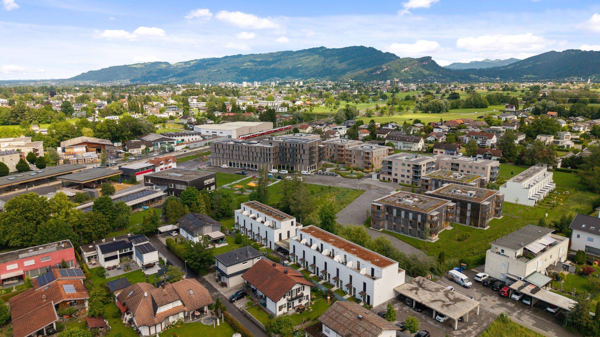 Weitläufige Luftaufnahme einer Wohnsiedlung mit Bergen im Hintergrund und viel Grün.