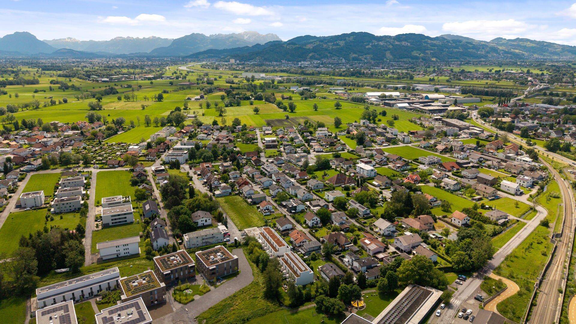Weitläufige Luftaufnahme einer Wohngegend mit grünen Wiesen und Bergen am Horizont unter blauem Himmel.