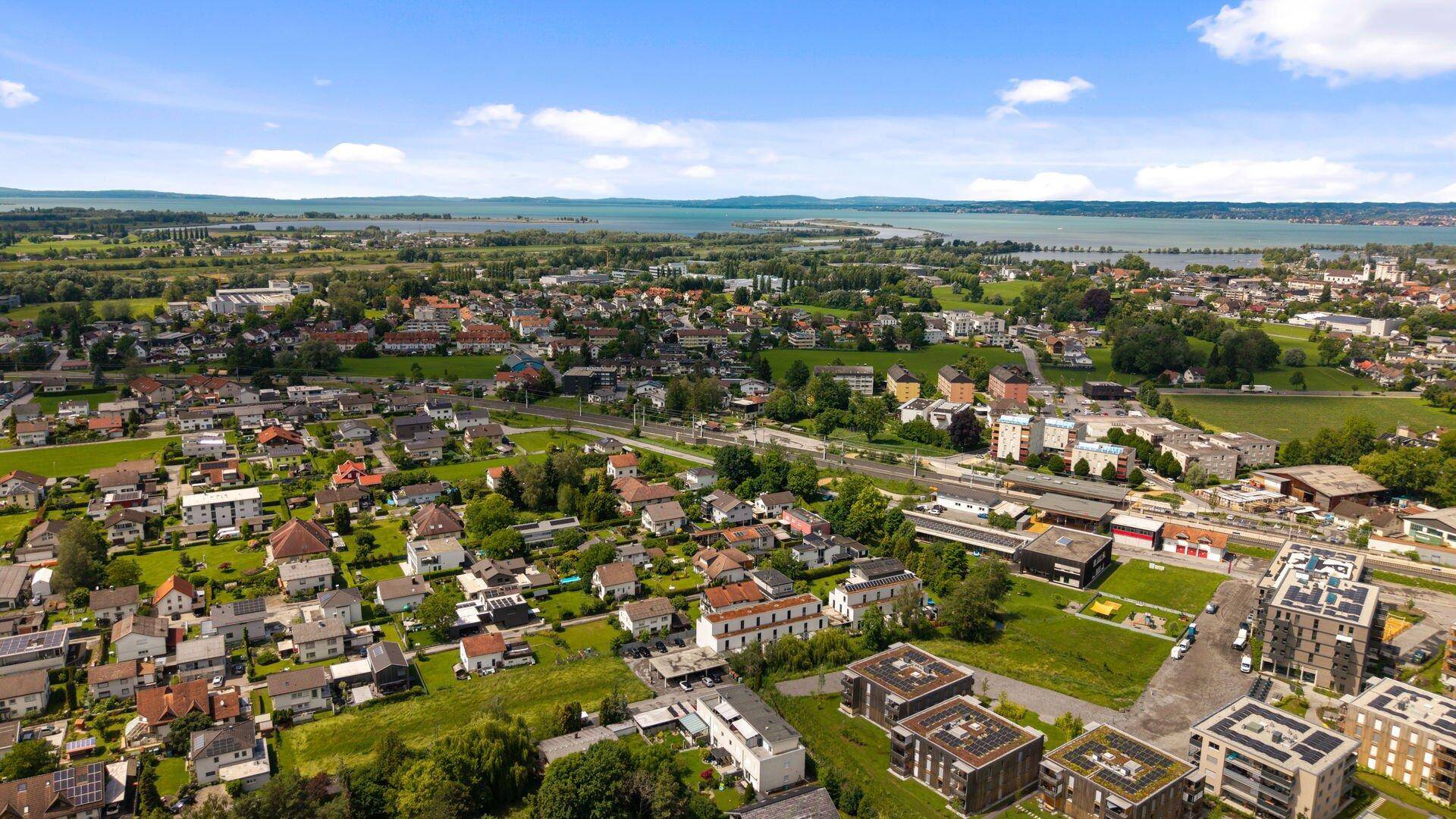 Luftaufnahme einer Wohnsiedlung mit grünen Feldern und einem See im Hintergrund unter blauem Himmel.