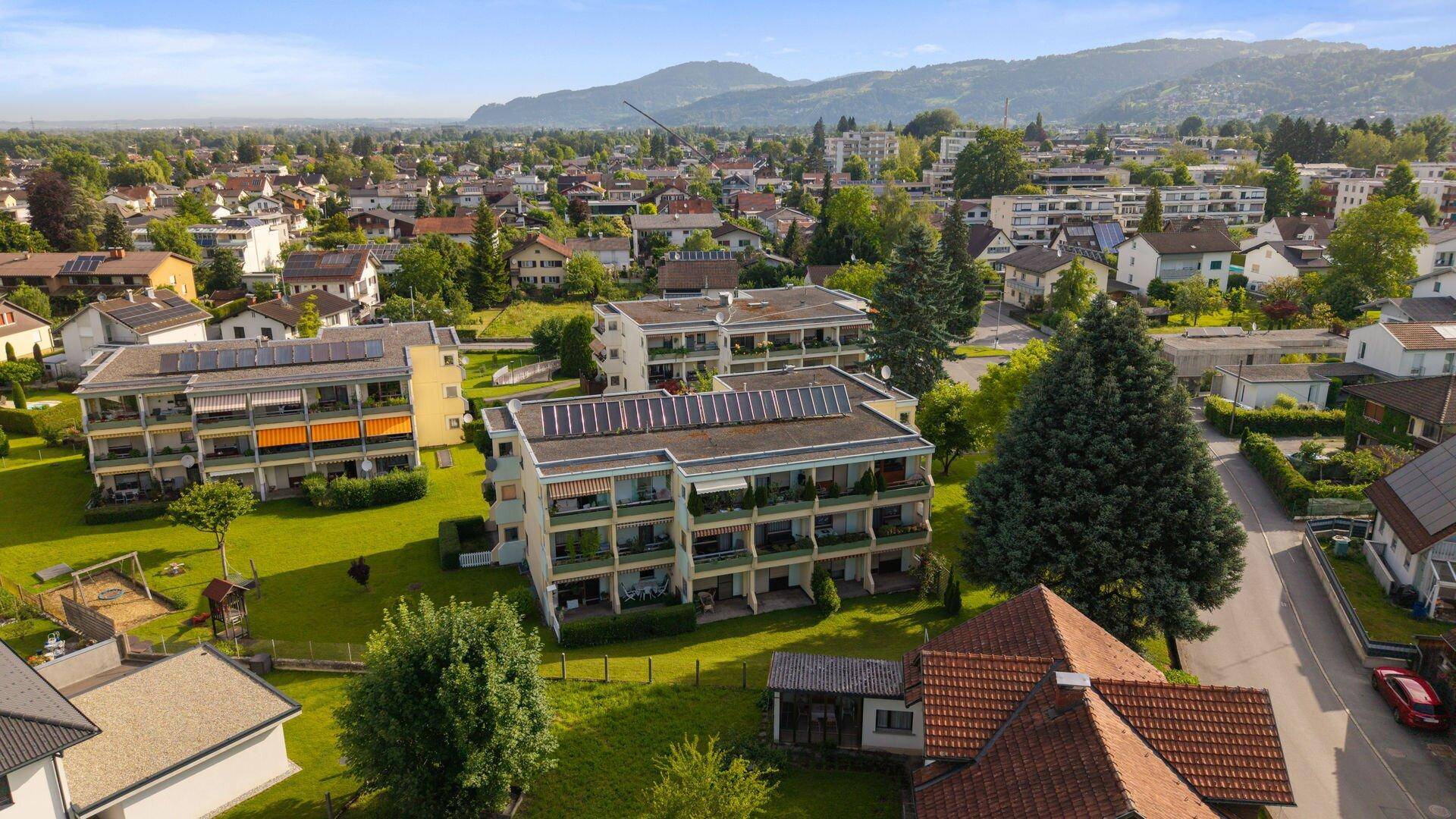 Luftaufnahme einer Wohnsiedlung mit Mehrfamilienhäusern, Grünflächen und Bergen im Hintergrund.