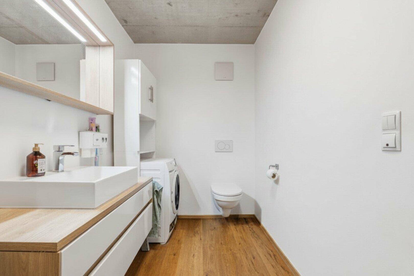 Funktionales Badezimmer mit WC, Waschbecken und integrierter Waschmaschine, schlicht und hell.