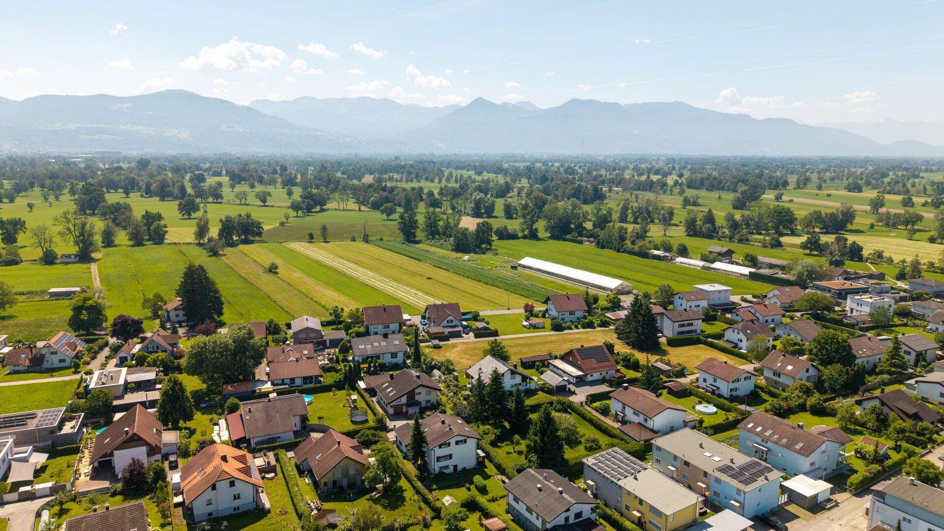 Luftaufnahme einer Wohnsiedlung angrenzend an Ackerland mit Bergblick unter blauem Himmel.