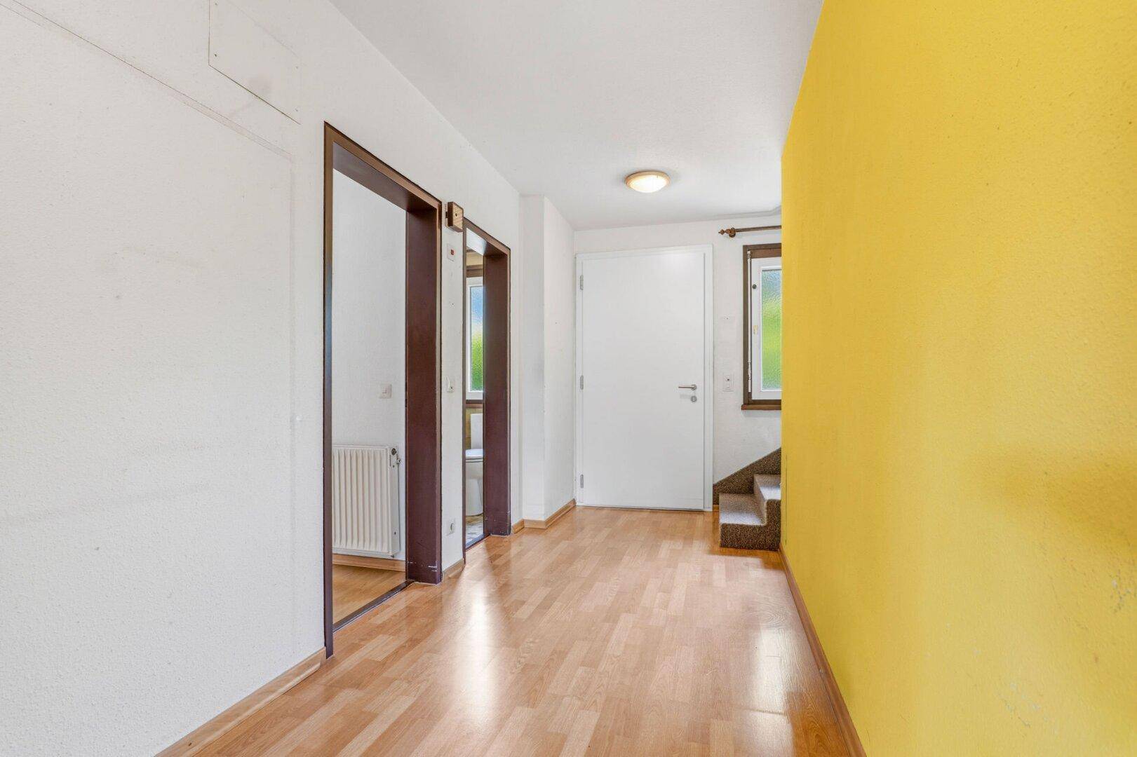Heller Flur mit gelber Akzentwand, Holzfußboden und Treppenaufgang.