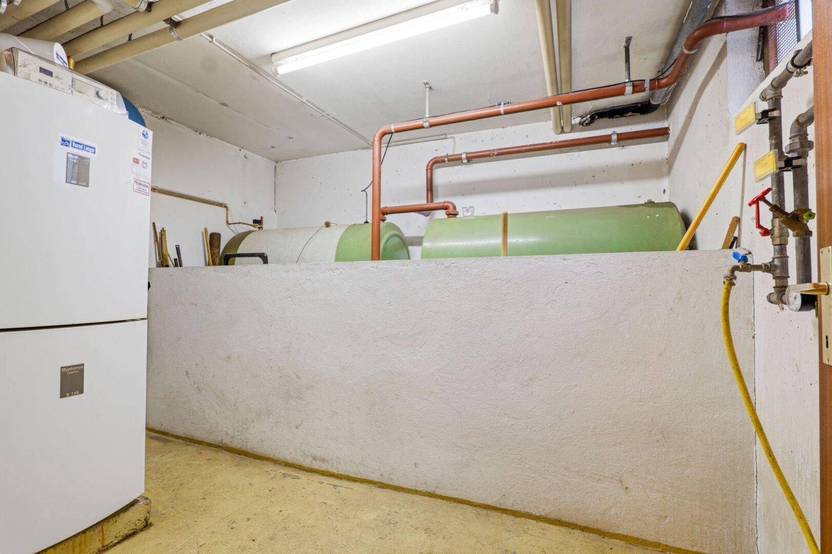 Technikraum im Keller mit Heizungsanlagen, großen Tanks und sichtbaren Rohrleitungen für die Versorgung.