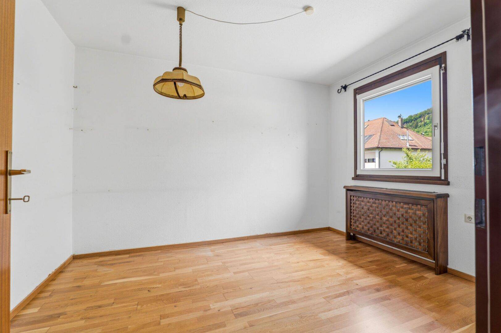 Schlafzimmer mit Parkettboden, Fenster und Blick auf die Dächer der Nachbarschaft.