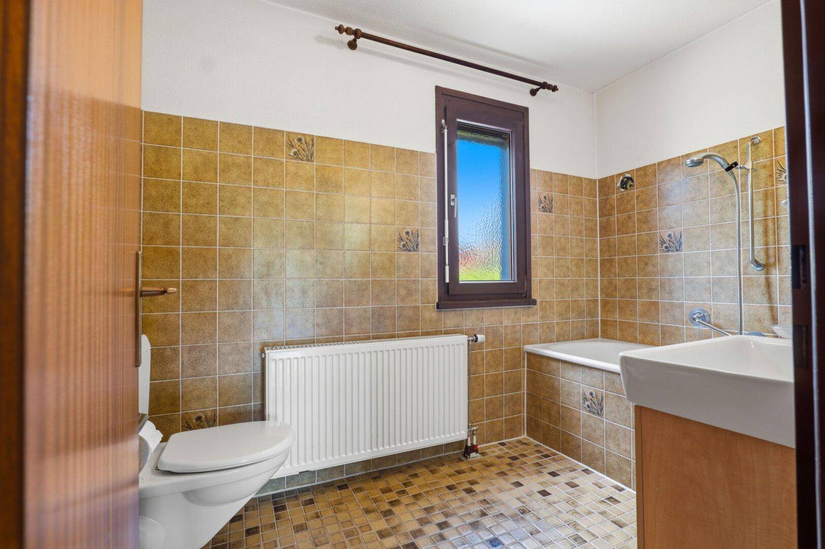 Badezimmer mit beigefarbenen Fliesen, Badewanne, WC und Fenster.
