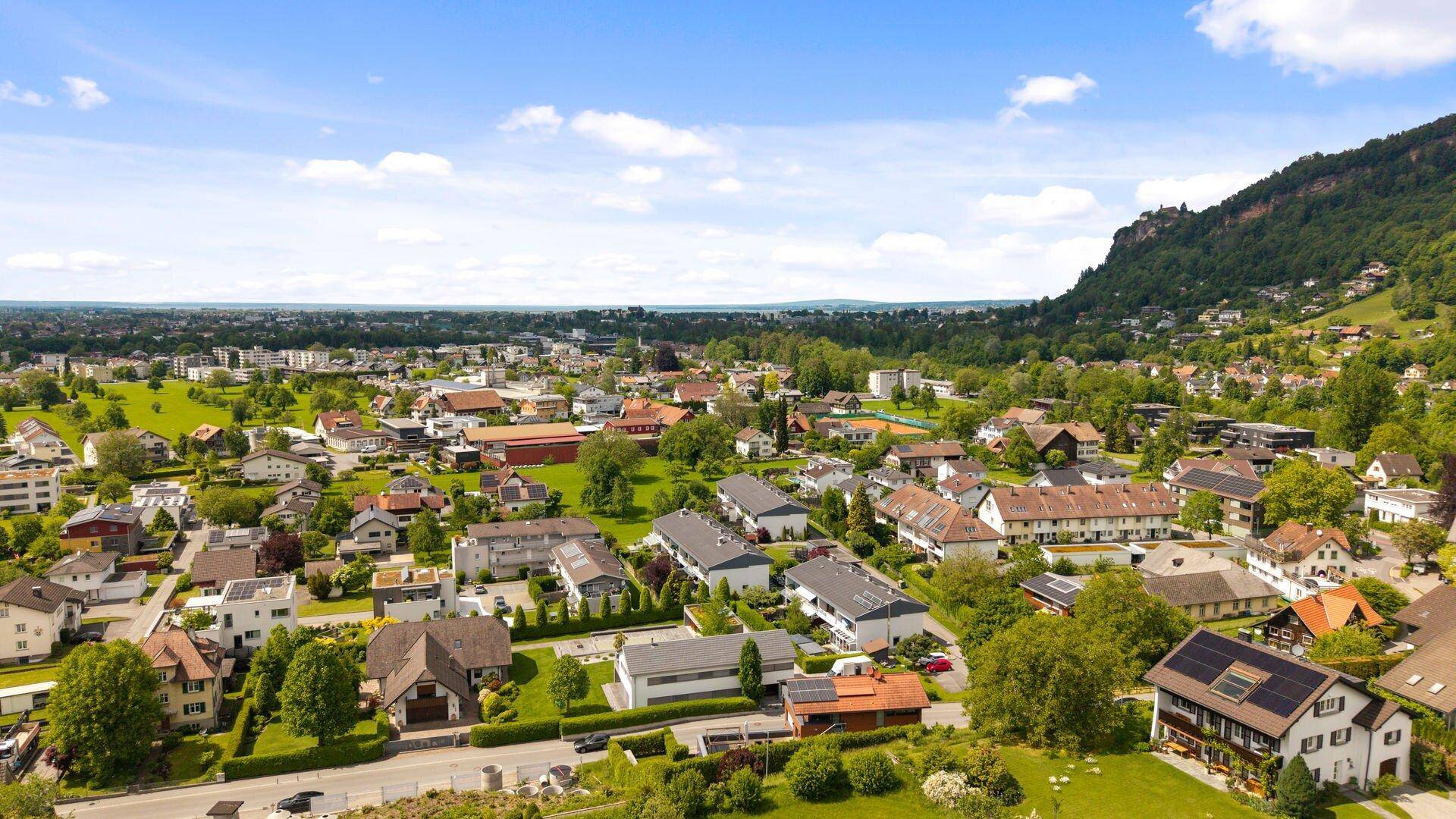 Weitläufige Luftaufnahme einer Siedlung mit vielen Häusern und grünen Gärten vor einer Bergkulisse.