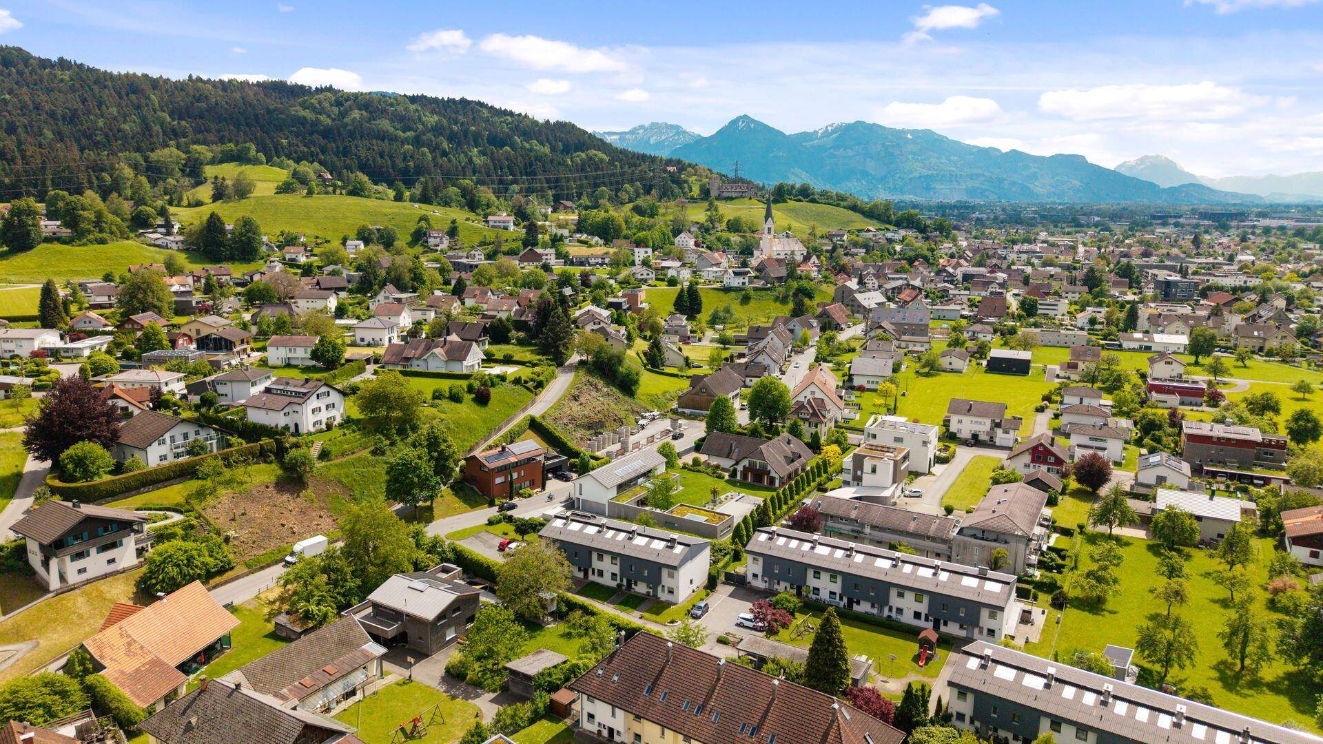 Weitläufige Luftaufnahme einer Siedlung mit grünen Landschaften und Bergen im Hintergrund unter blauem Himmel.