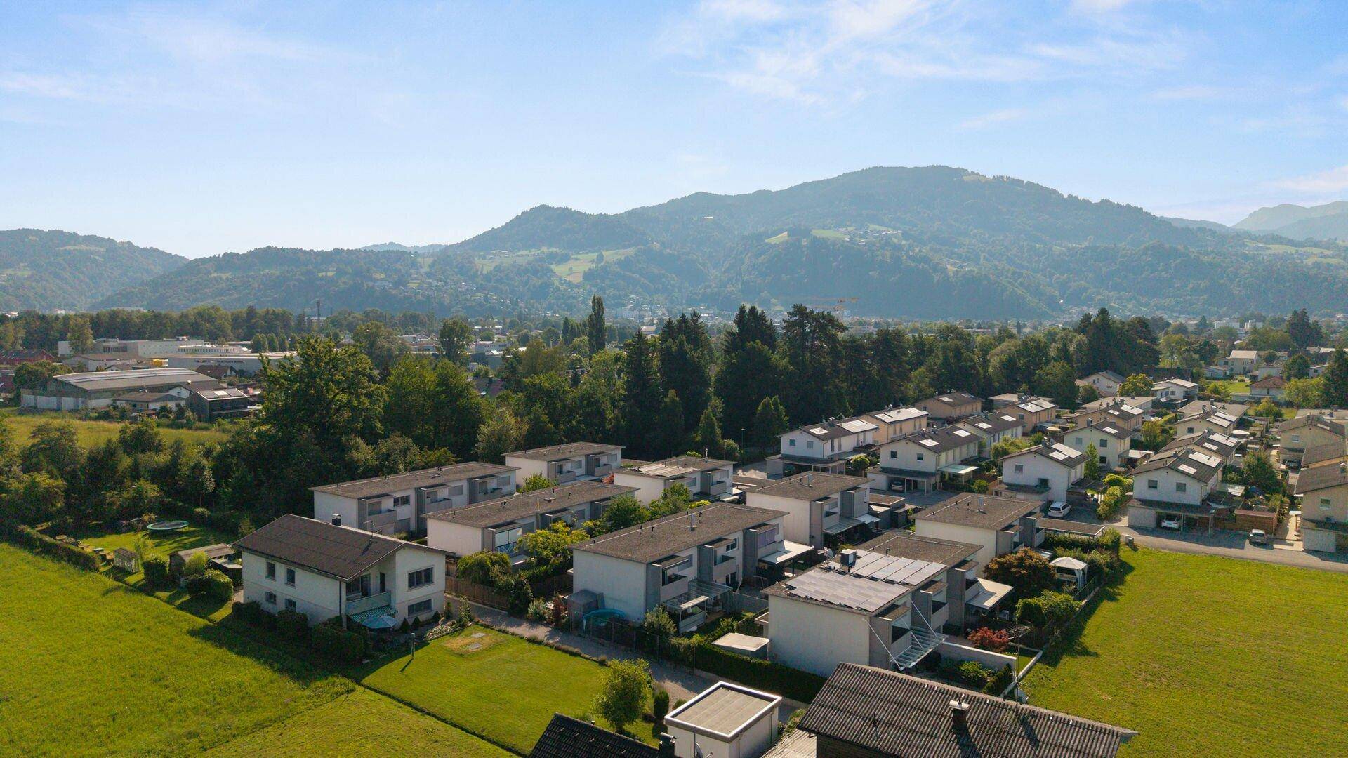 Weitläufige Luftaufnahme einer Wohnsiedlung mit grünen Feldern und Bergen im Hintergrund.
