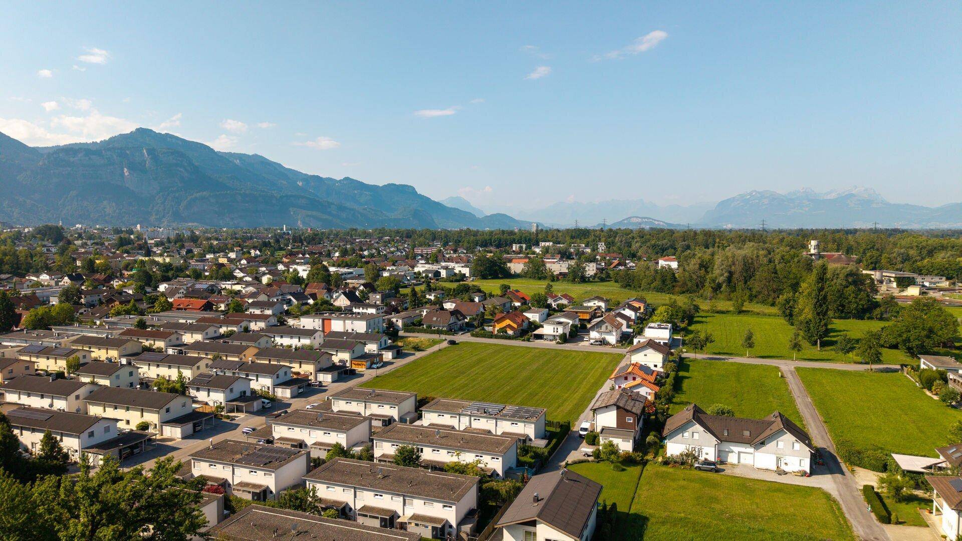 Luftaufnahme einer Wohnsiedlung mit grünen Wiesen und Bergen am Horizont.