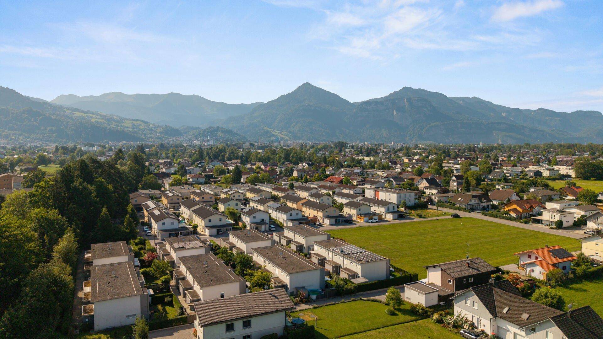 Panoramablick auf eine Wohnsiedlung mit modernen Häusern und weiten Grünflächen, umgeben von Bergen.