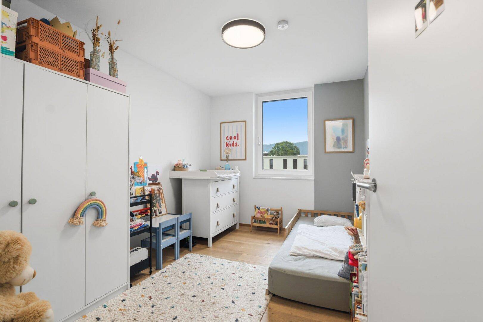 Helles Kinderzimmer mit Holzboden, großem Fenster und Blick ins Grüne.