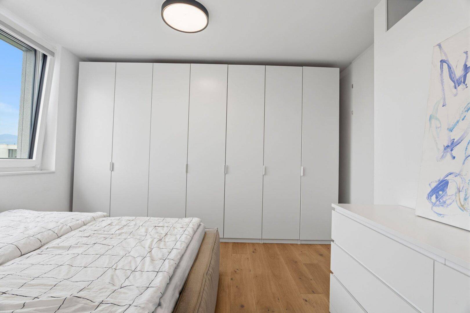 Helles Schlafzimmer mit Einbauschränken, Holzboden und großem Fenster.