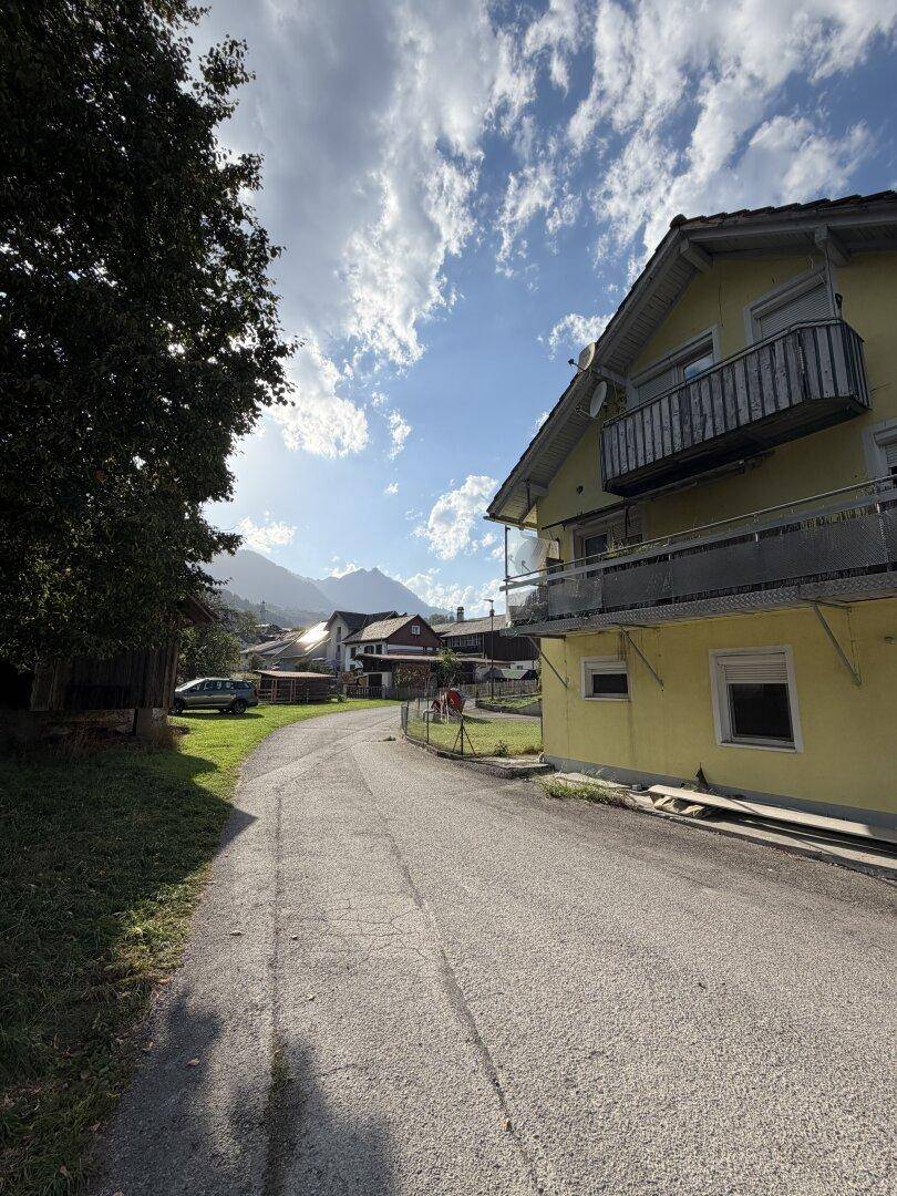 Gelbes Mehrfamilienhaus in ländlicher Umgebung mit Balkonen und Bergblick unter blauem Himmel.