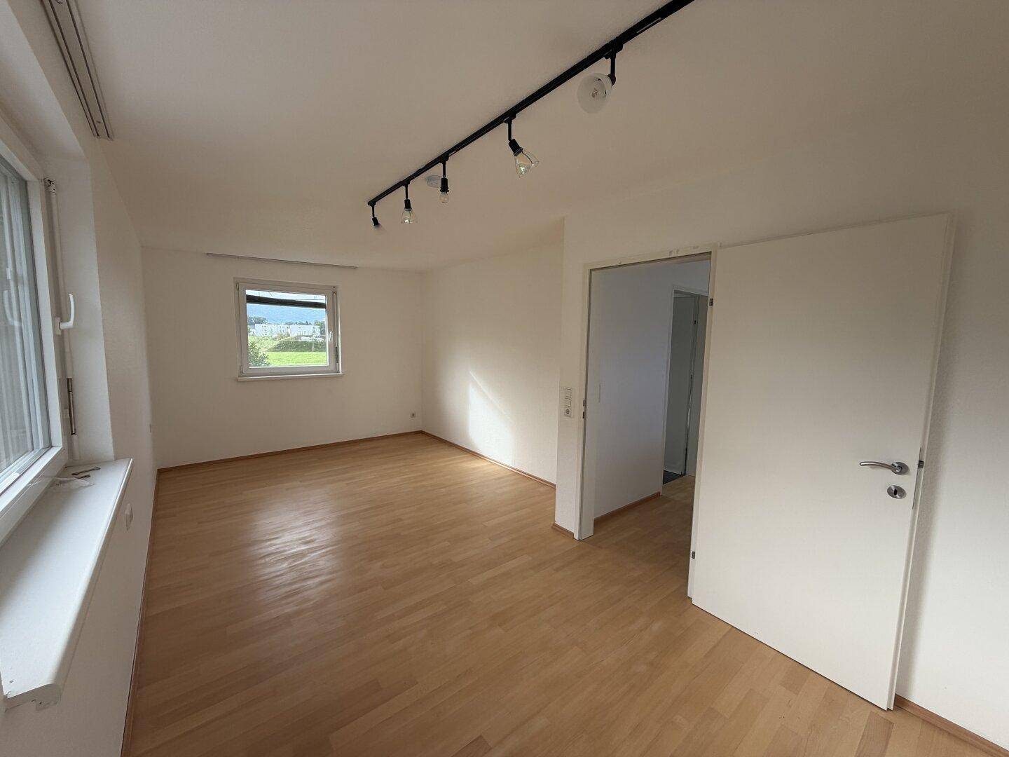 Helles Zimmer mit Holzboden, zwei Fenstern und moderner Deckenbeleuchtung.