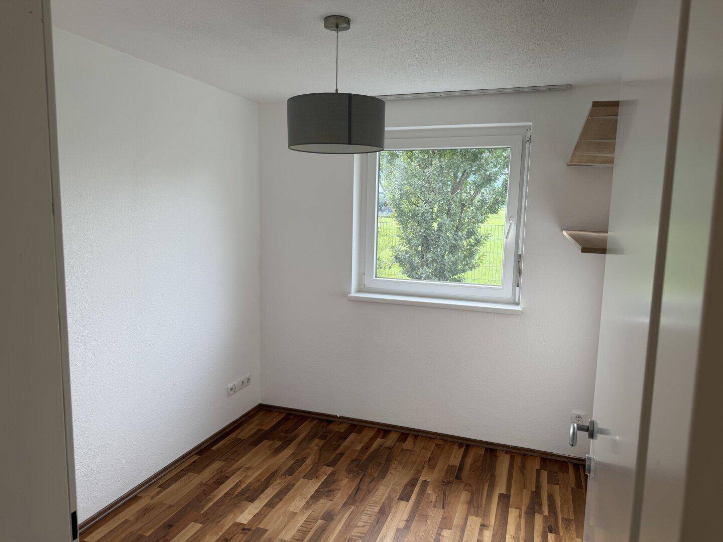 Kleines helles Zimmer mit Holzboden und Fenster mit Blick ins Grüne.