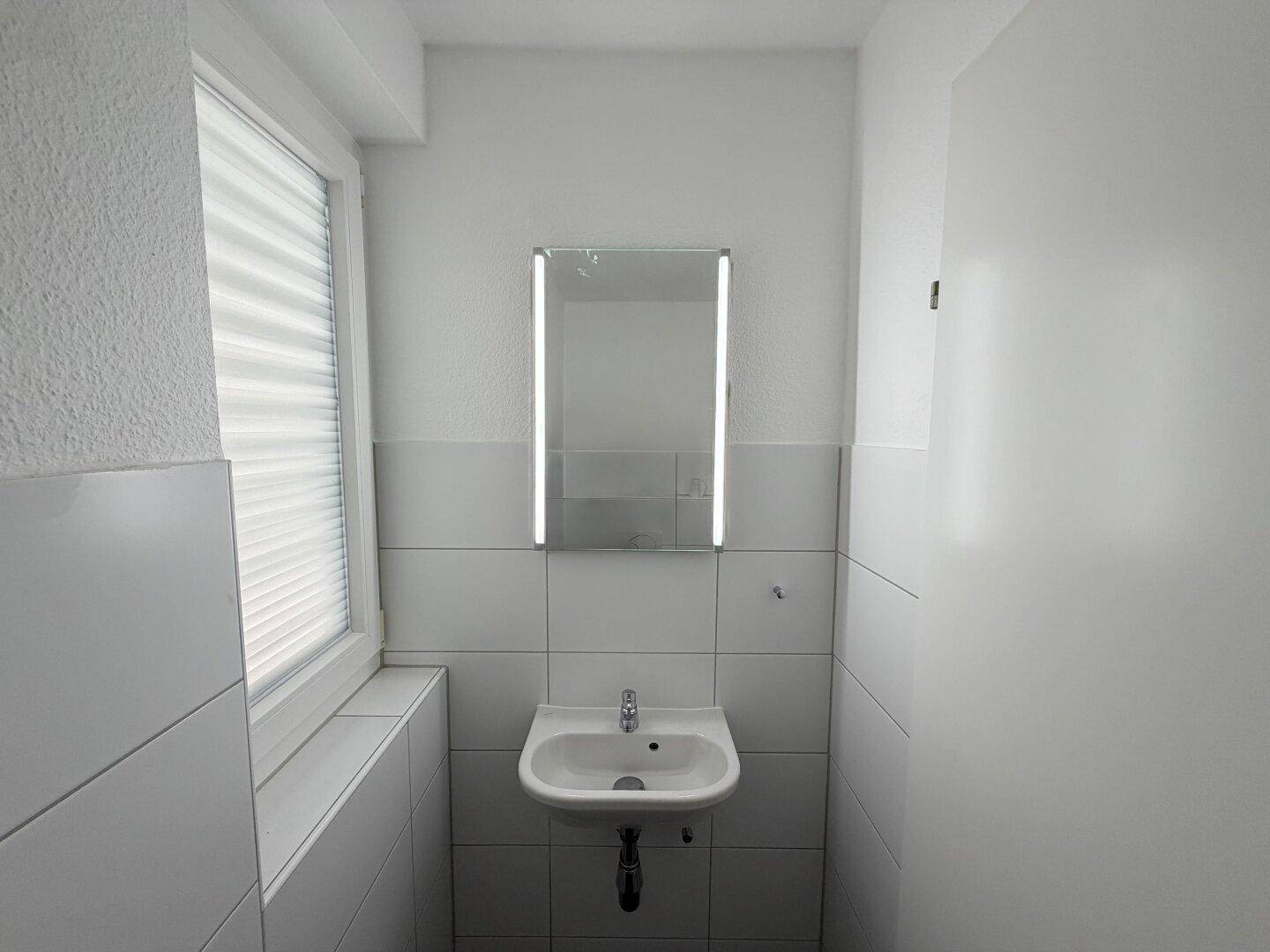 Helles Gäste-WC mit Einzelwaschbecken, beleuchtetem Spiegel und Fenster.