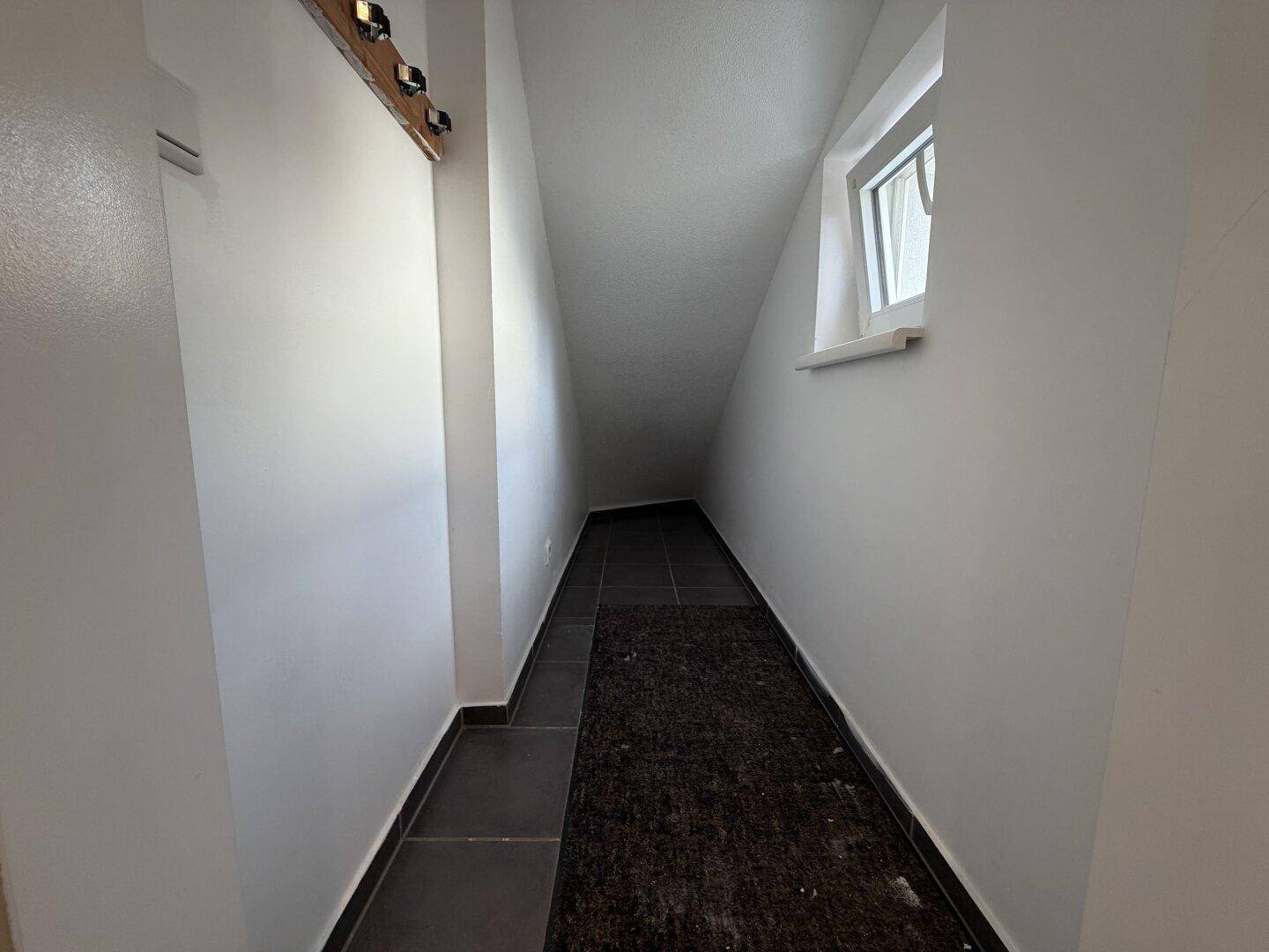 Schmaler Gang mit Dachschräge, dunklen Fliesen und einem Fenster am Ende.