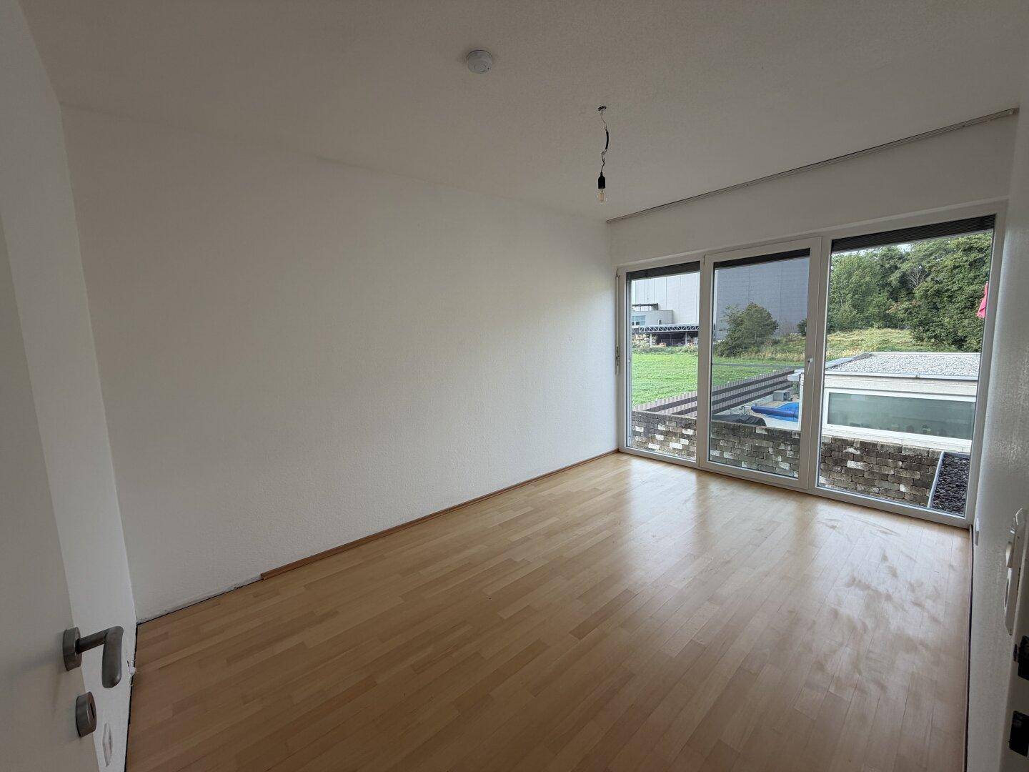 Geräumiges Zimmer mit Holzboden und großen Fenstern mit Blick ins Grüne.