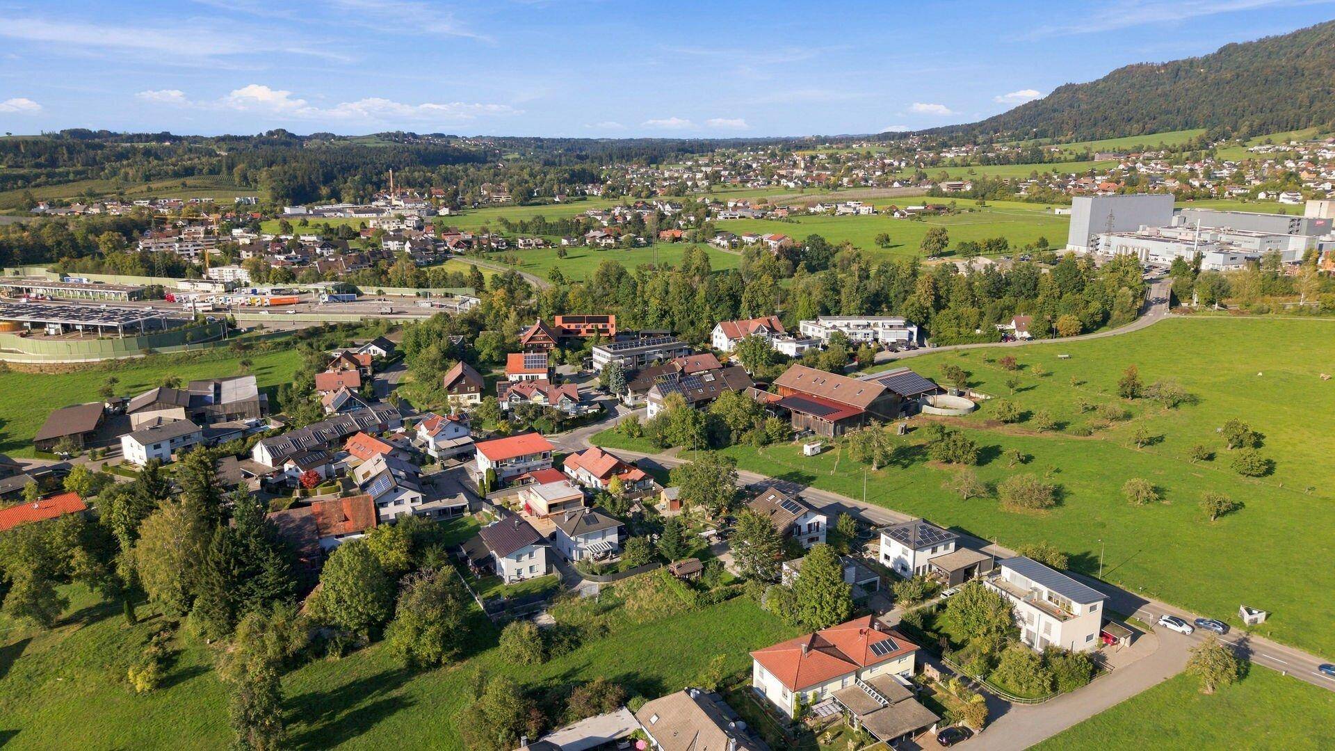 Weitläufige Luftaufnahme einer ländlichen Siedlung, umgeben von grünen Feldern und bewaldeten Hügeln.