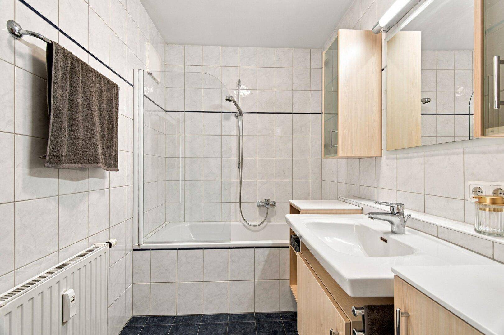 Helles Badezimmer mit Badewanne, Duschabtrennung, Waschbecken und Heizkörper.