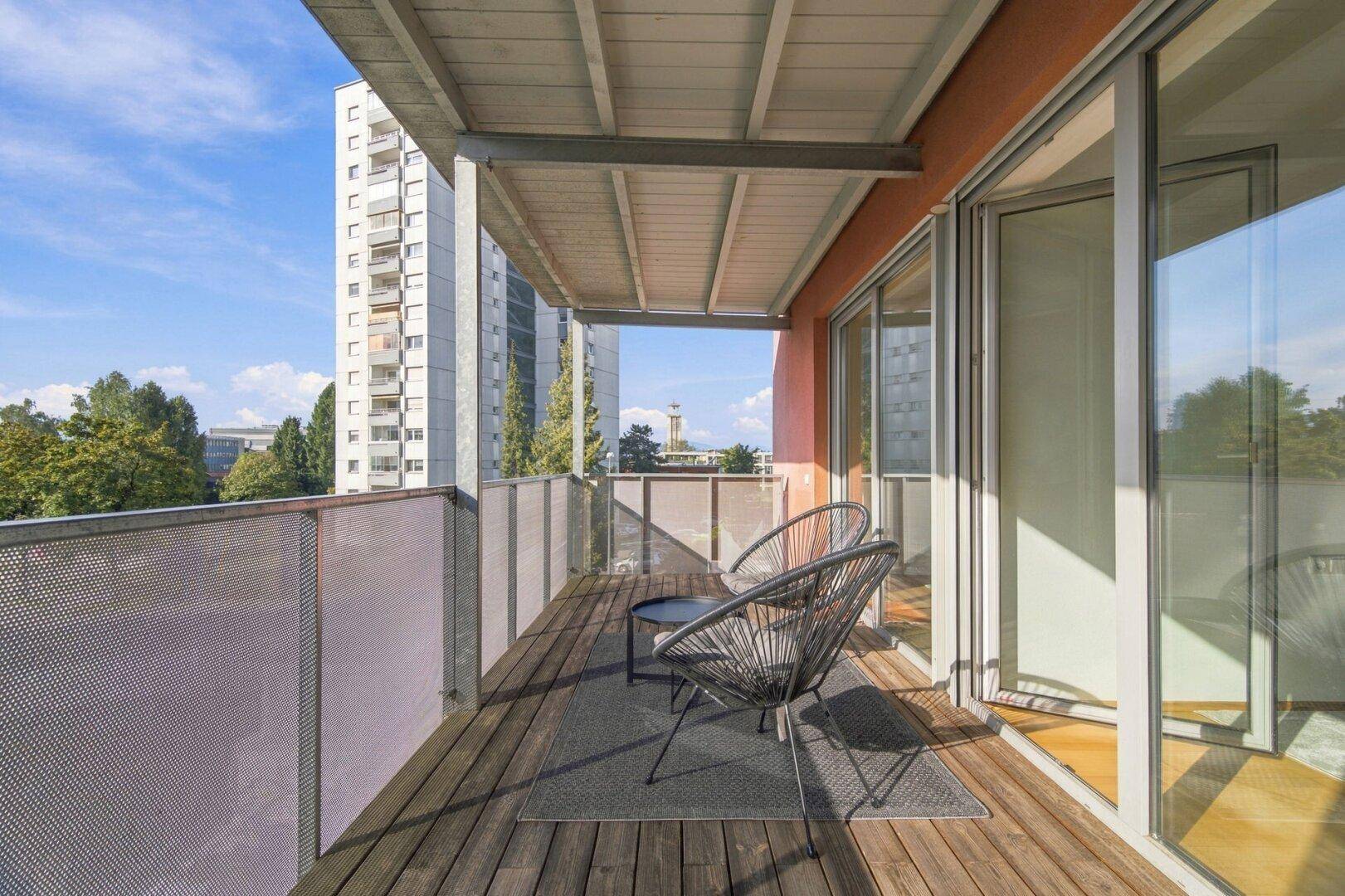 Geräumiger Balkon mit Holzfußboden, Sitzgelegenheiten und Blick auf die umliegenden Gebäude.