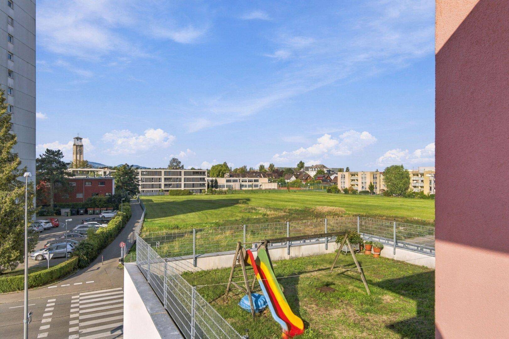 Blick vom Balkon auf einen grünen Bereich mit Spielplatz und umliegenden Gebäuden.