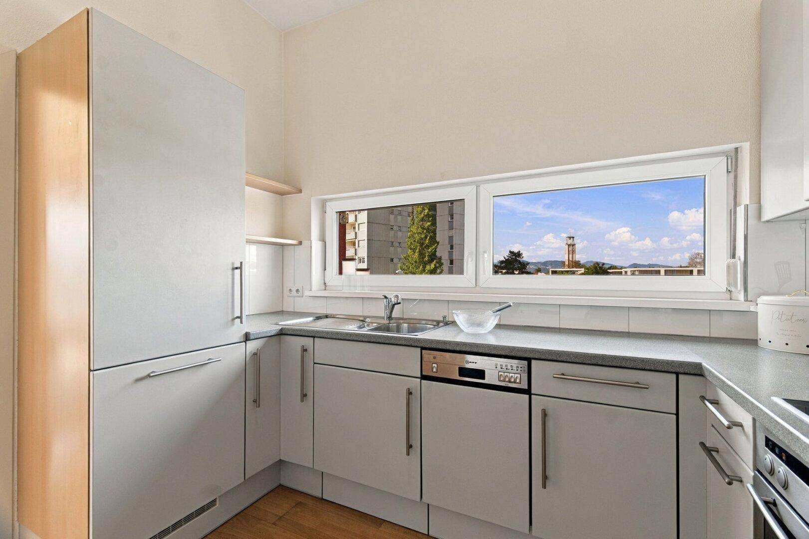 Helle Küche mit grauen Schränken, Arbeitsplatte und Fenster mit Blick auf die Umgebung.