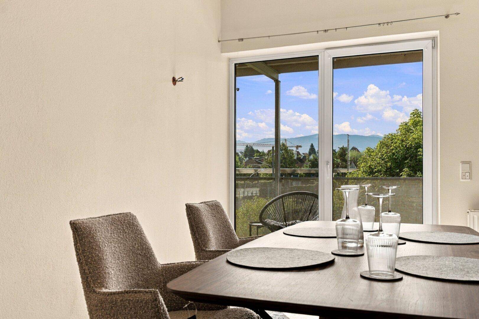 Nahaufnahme des Esstisches mit Blick durch die Balkontür auf die umliegende Landschaft und Berge.
