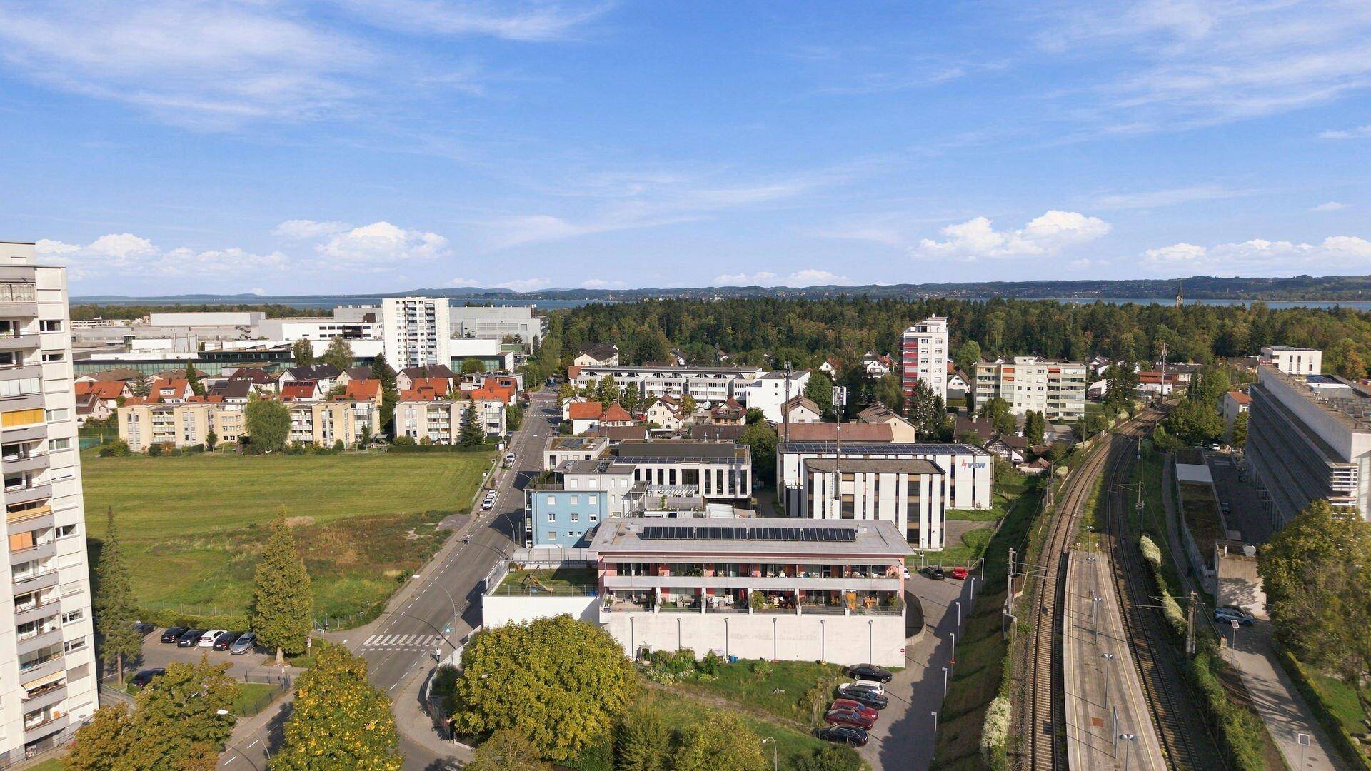 Weitläufige Luftaufnahme eines Stadtteils mit Wohngebäuden, Grünflächen und einem See in der Ferne.