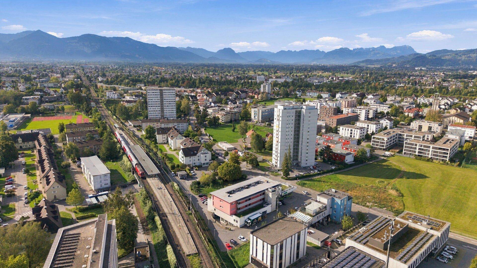 Panoramische Luftaufnahme einer Stadtlandschaft mit Bergen im Hintergrund und einer Zugstrecke.