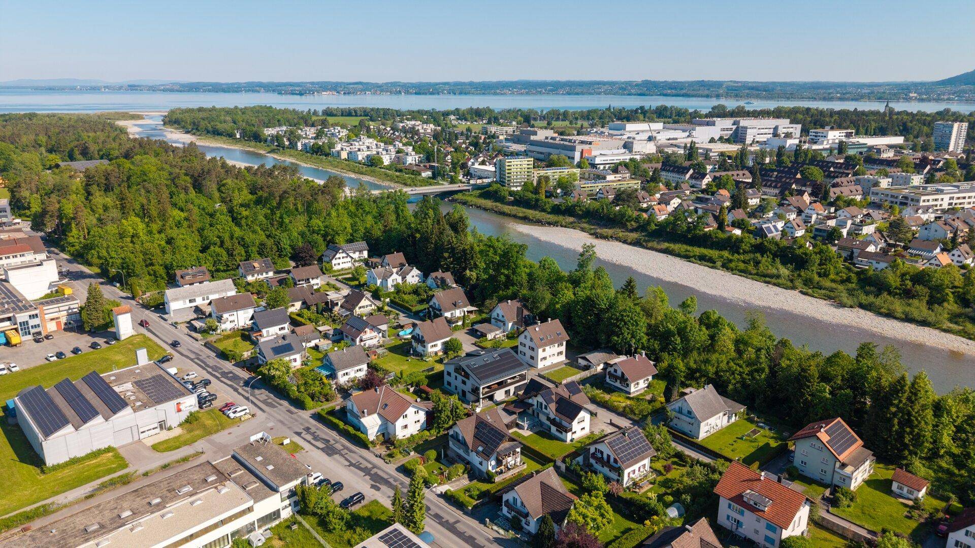 Weitläufige Luftaufnahme einer Stadt am Flussufer mit einem See im Hintergrund und Wohngebieten.