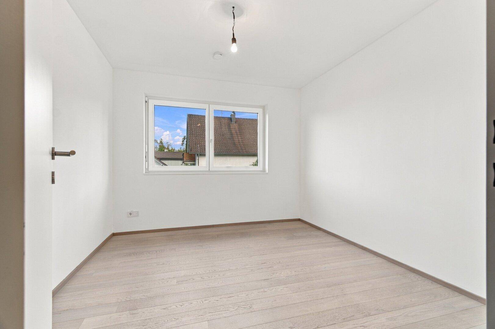 Unmöbliertes Schlafzimmer mit hellem Holzboden und Fenster mit Blick auf die Dächer.