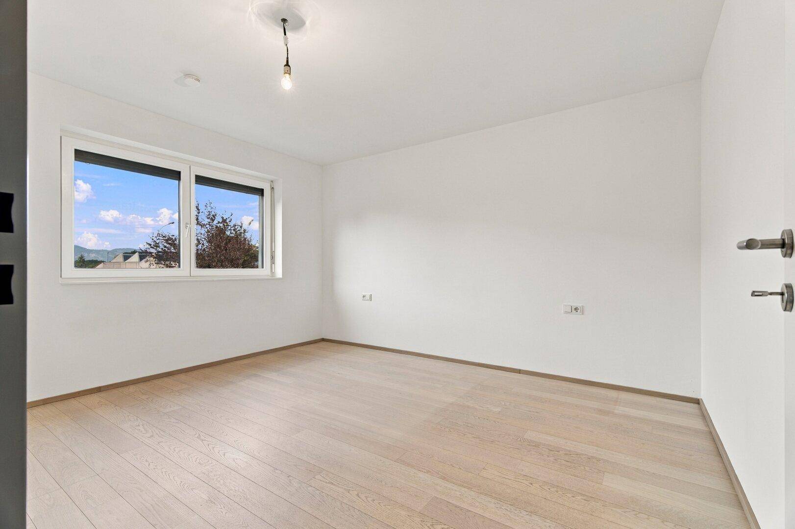 Unmöbliertes Schlafzimmer mit hellem Holzboden und großem Fenster mit Blick ins Grüne.