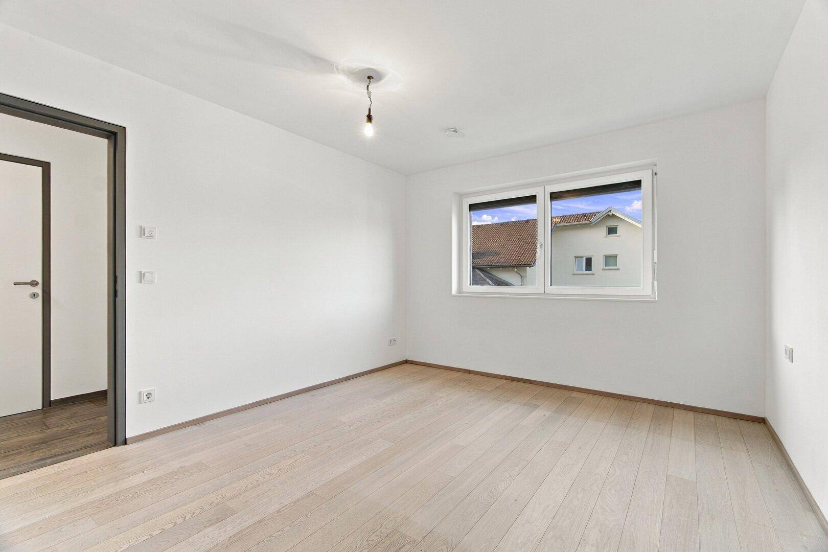 Helles, unmöbliertes Schlafzimmer mit Holzboden und Fensterblick auf die Nachbarschaft.