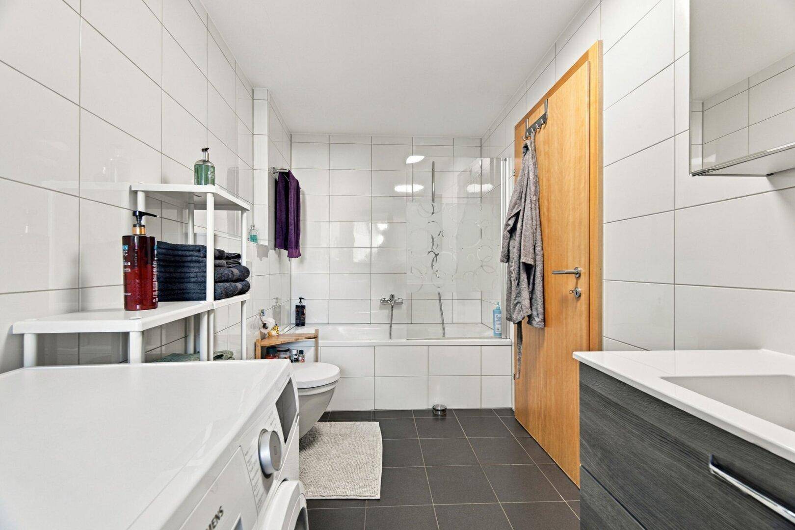 Modernes Badezimmer mit weißen Fliesen, Badewanne und Waschmaschine.
