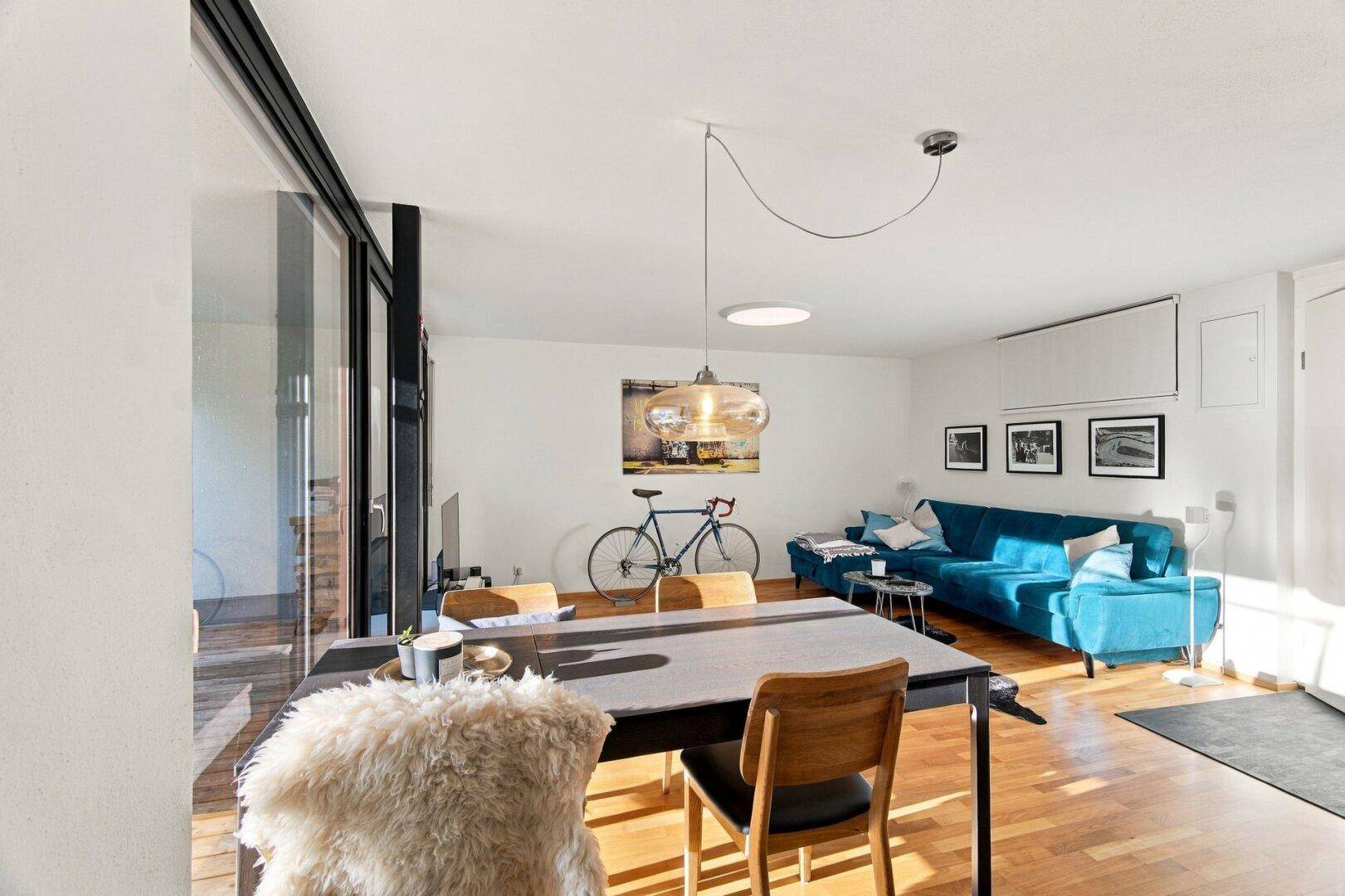 Heller Wohn- und Essbereich mit blauer Couch, Esstisch und großen Fenstern.