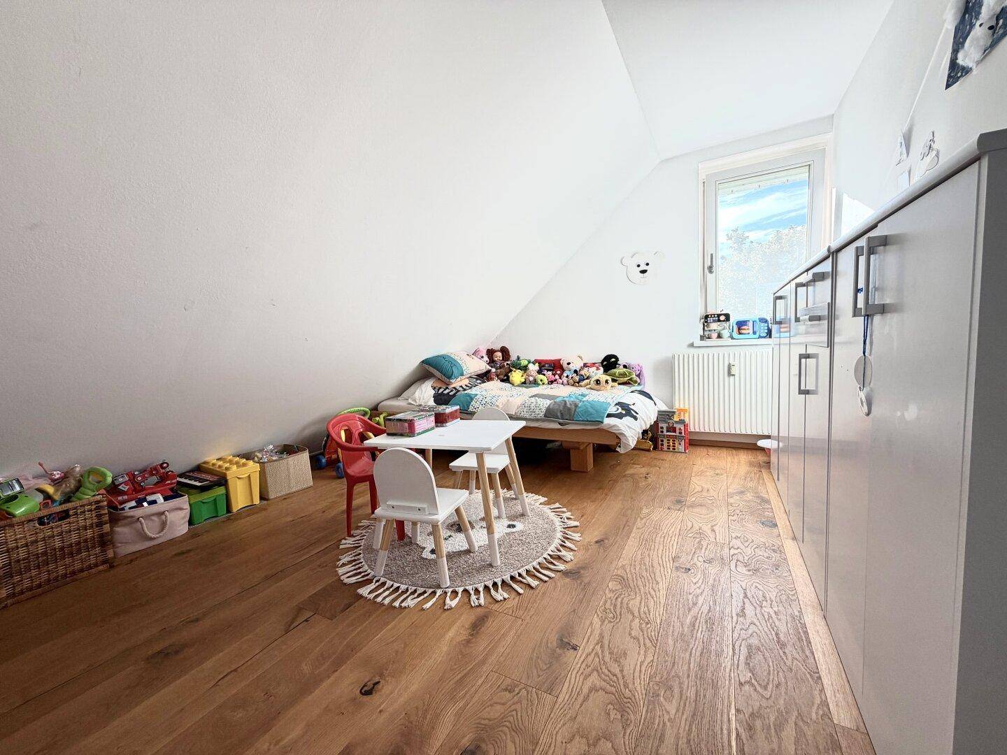 Helles Kinderzimmer mit Holzboden, Fenster und Spielzeug, gemütlich unter der Dachschräge gelegen.