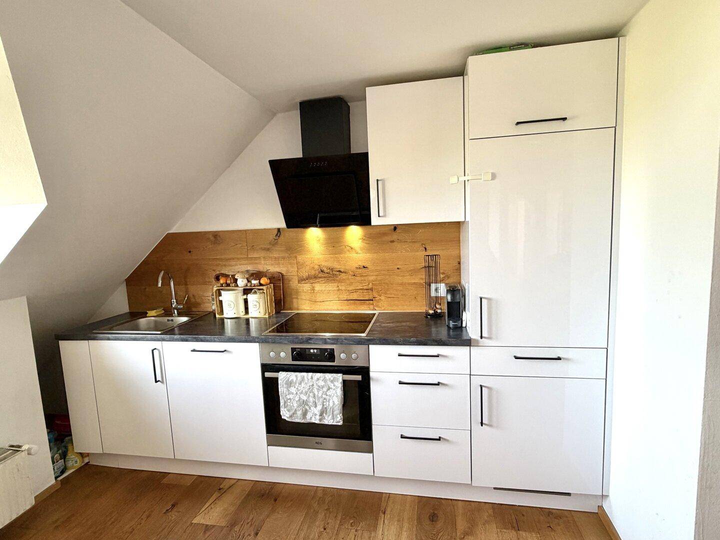 Moderne Einbauküche mit weißen Fronten, dunkler Arbeitsplatte, Holzwandverkleidung und integrierten Geräten unter der Dachschräge.