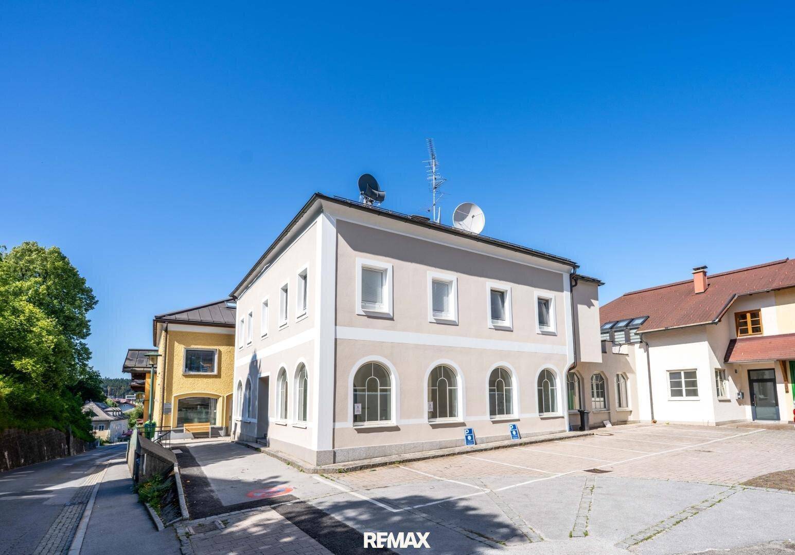 Gebäudeansicht mit heller Fassade, Bogenfenstern und Parkplätzen vor dem Eingangsbereich.