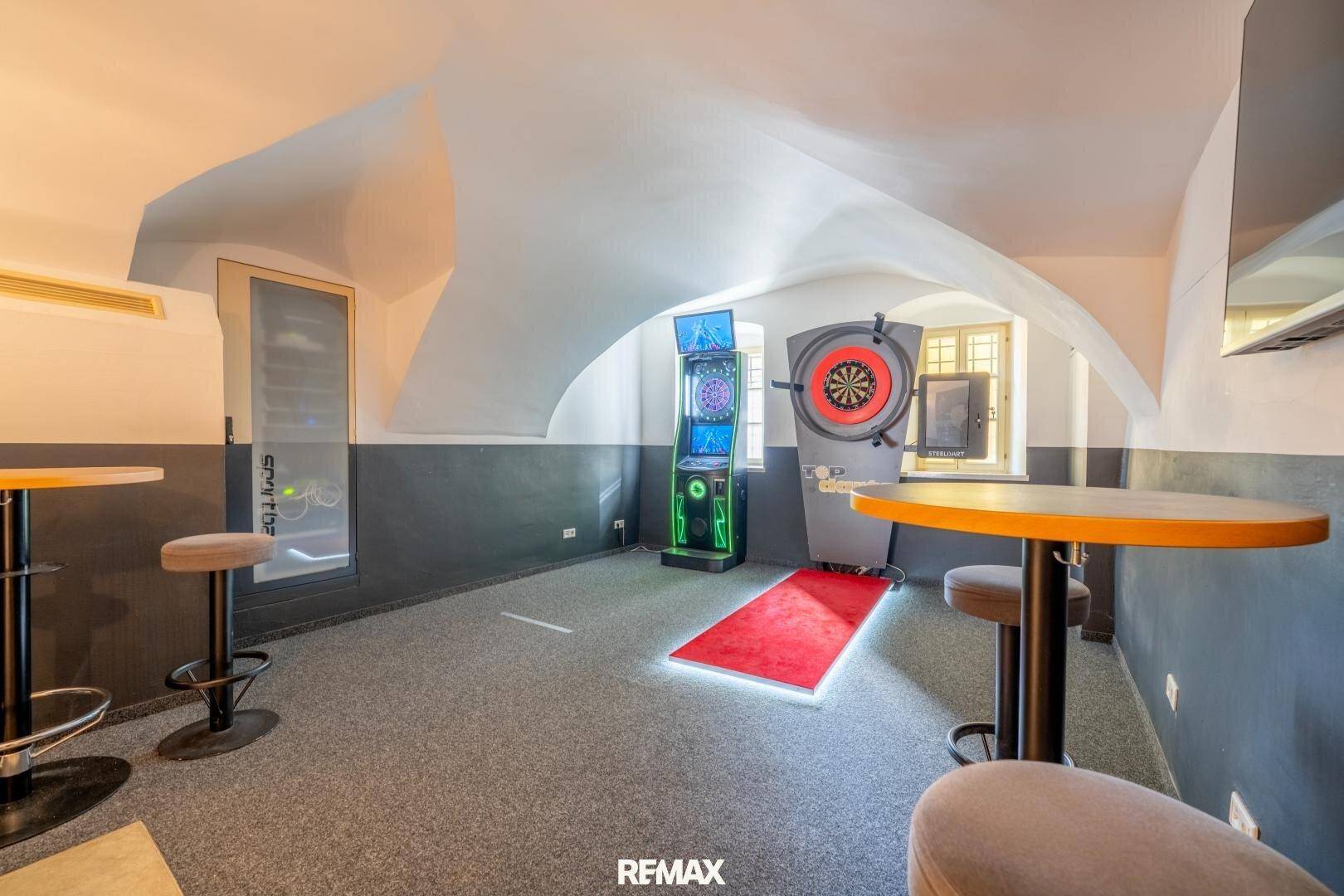 Spielzimmer mit Dartscheibe, Stehtischen und Teppichboden unter gewölbter Decke.