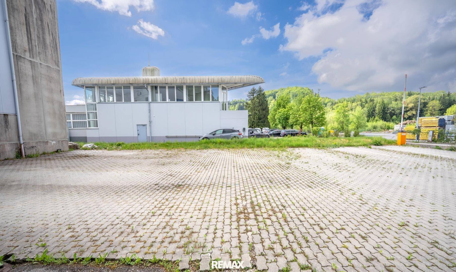 Großer gepflasterter Parkplatz mit einem weiteren Gebäude im Hintergrund und grüner Umgebung.