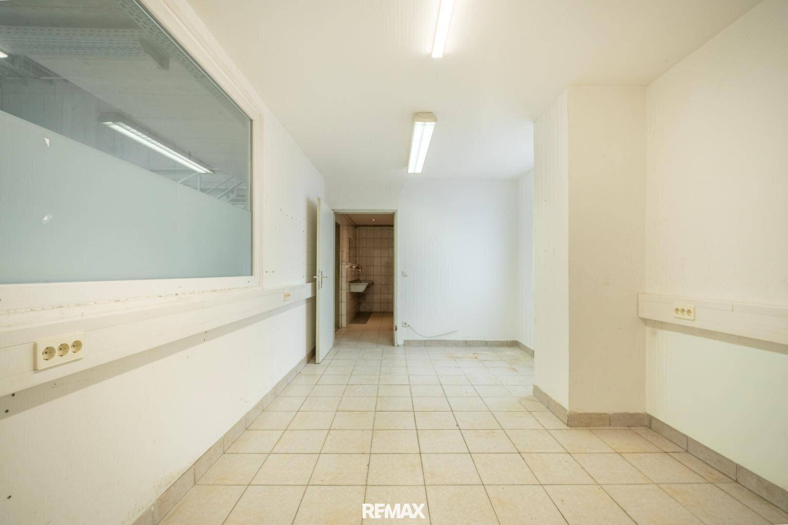 Einfacher Büroraum mit Fliesenboden, weißen Wänden und einem Fenster zur Halle.