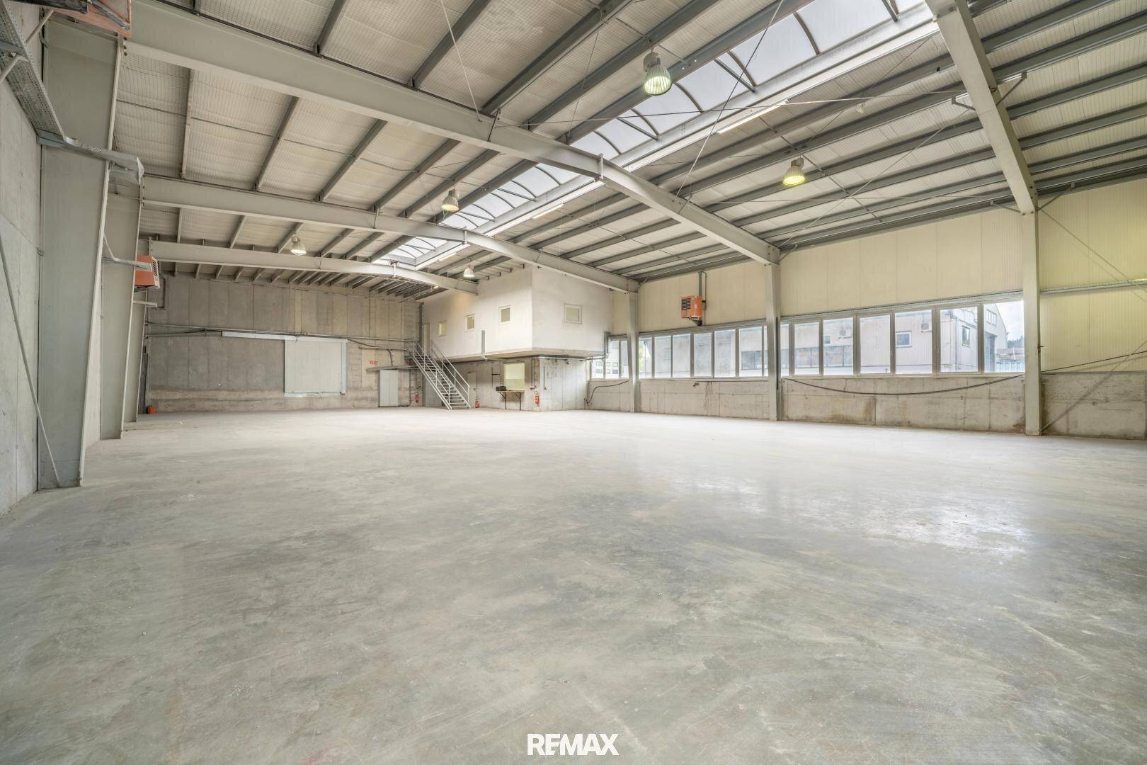 Große, helle Lagerhalle mit hohen Decken, Betonboden und einer erhöhten Bürofläche im Hintergrund.